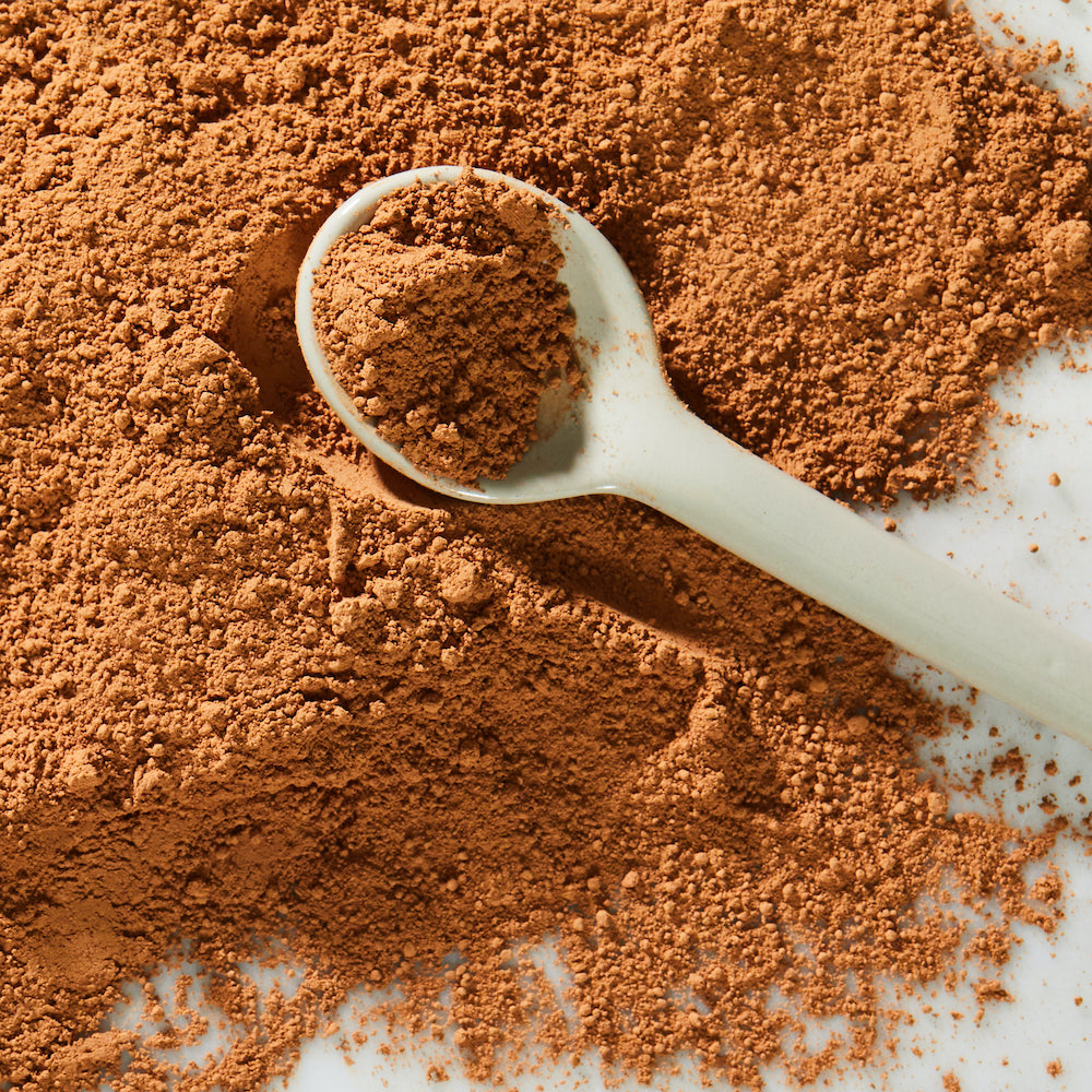 A white measuring spoon cups a portion of light brown cocoa powder from a pile of cocoa powder spilled out onto a bright marble surface.