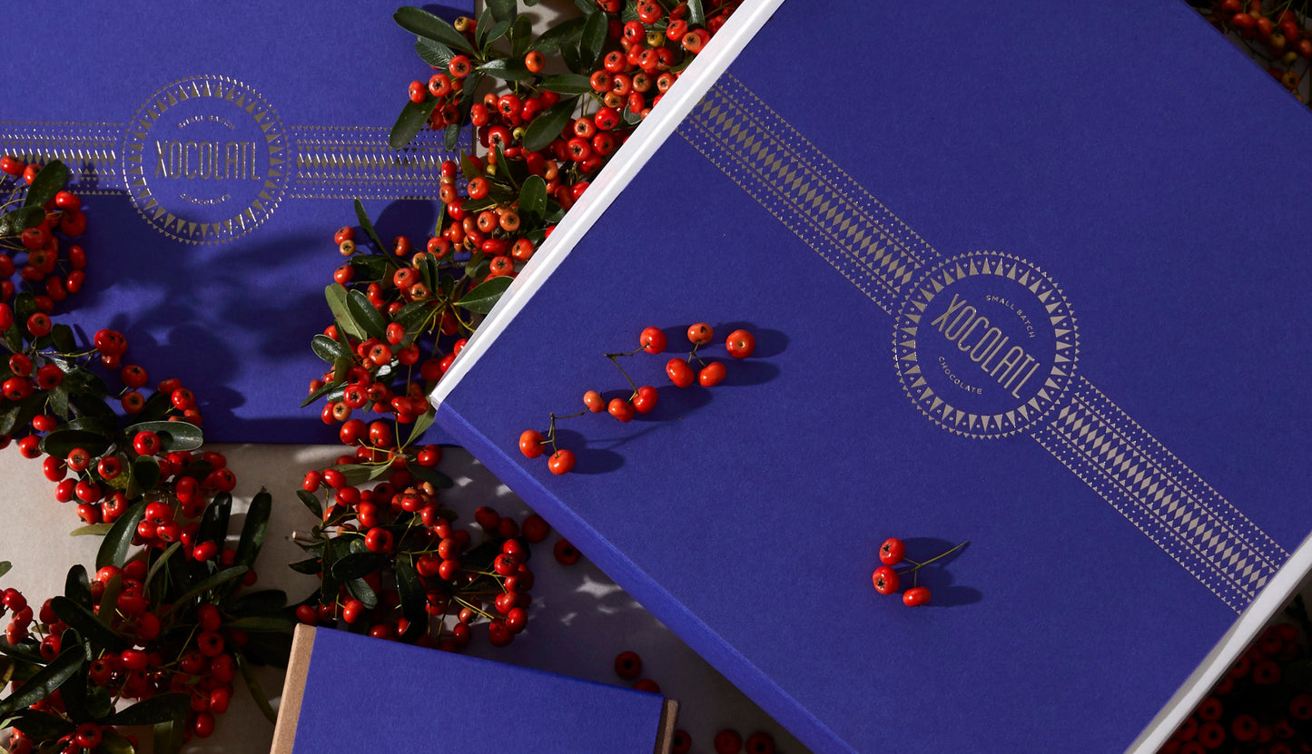 Three royal blue gift boxes (small, medium & large) with the Xocolatl logo stamped in silver foil are spread on a table covered in festive red berries & greenery.