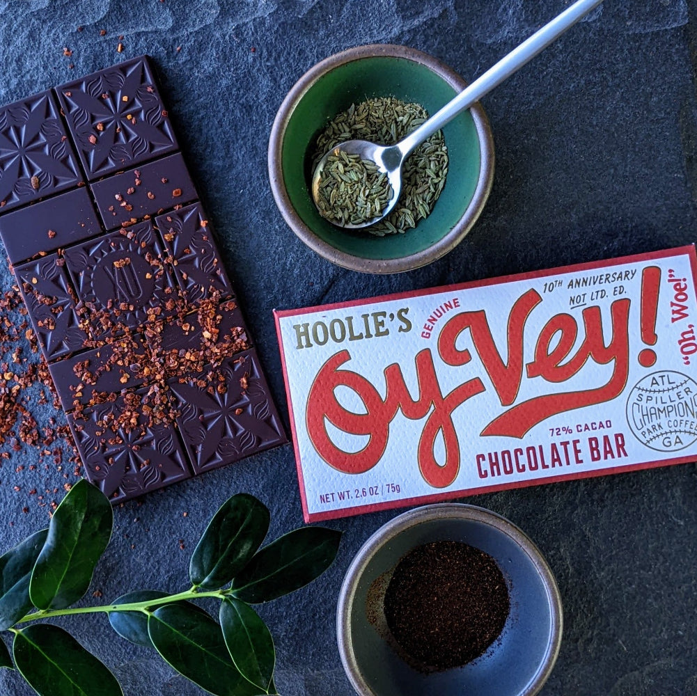 Xocolatl dark chocolate bar on a dark surface with dried chili peppers, toasted fennel & sumac spices beside the bar wrapper which is called Oy Vey.