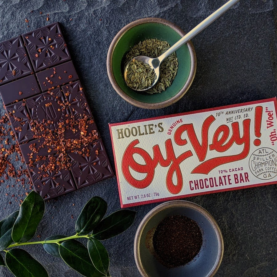 Xocolatl dark chocolate bar on a dark surface with dried chili peppers, toasted fennel & sumac spices beside the bar wrapper which is called Oy Vey.