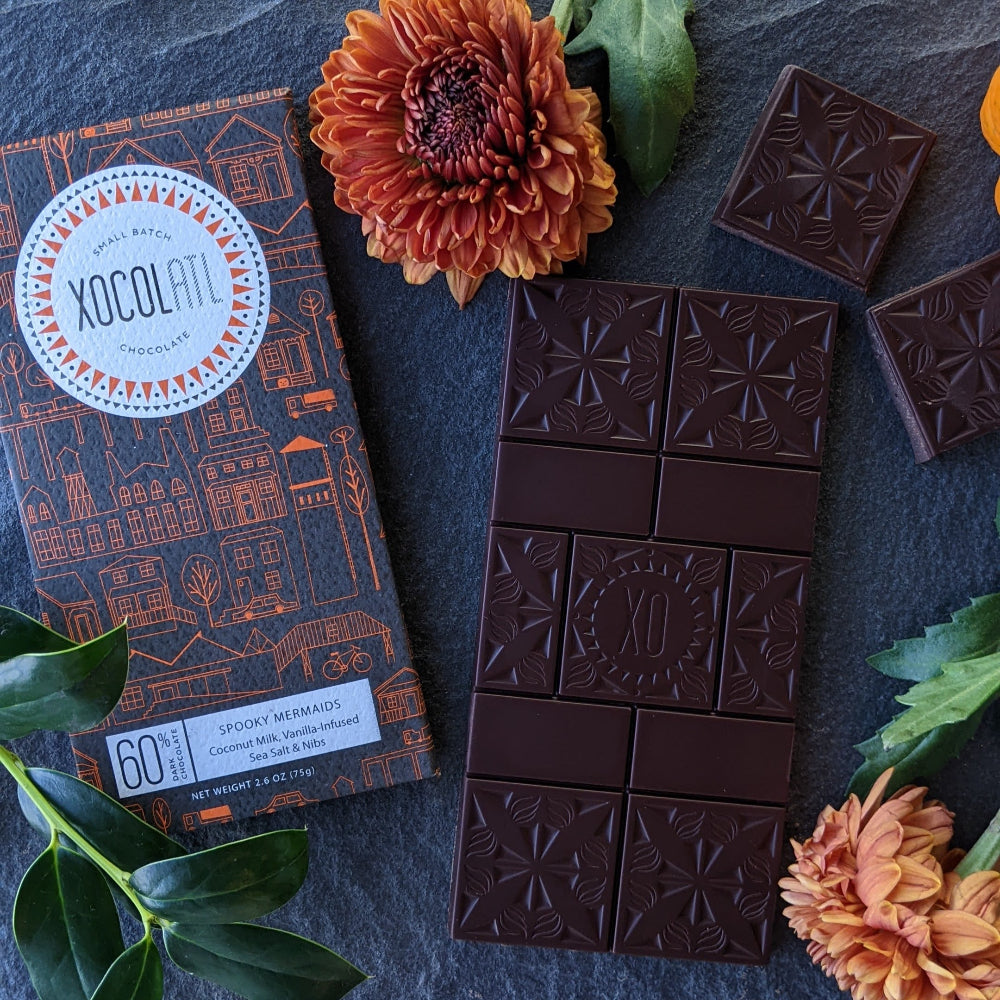 Xocolatl Chocolate bar wrapped in a Halloween-themed black and orange wrapper with unwrapped chocolate bar and orange marigold flowers.