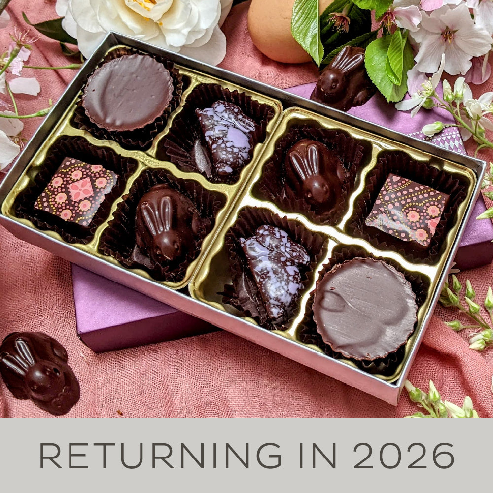 An 8 piece box of chocolate bonbons opened to show a variety of bunny-shaped bonbons sitting with white and pink spring flowers.A banner along the bottom reads "Returning in 2026".