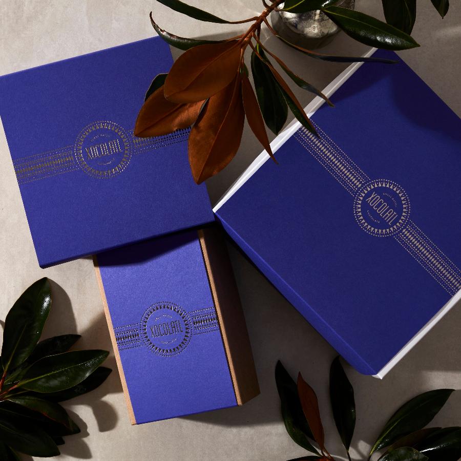 3 royal blue Xocolatl Chocolate gift boxes in royal blue with the circular Xocolatl logo stamped in silver in the center of each box. There are decorative magnolia branches alongside the boxes.