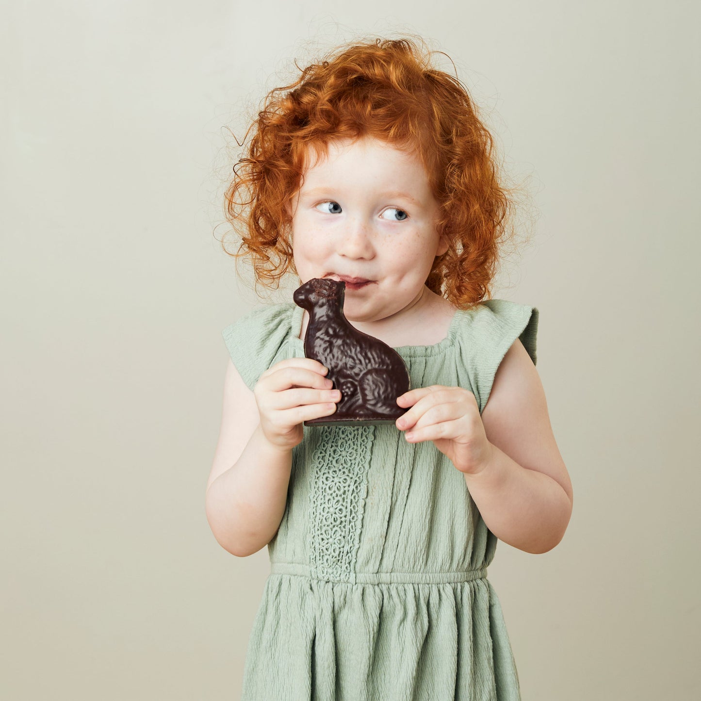 A little girl holding an earless chocolate Easter bunny smiles mischievously.