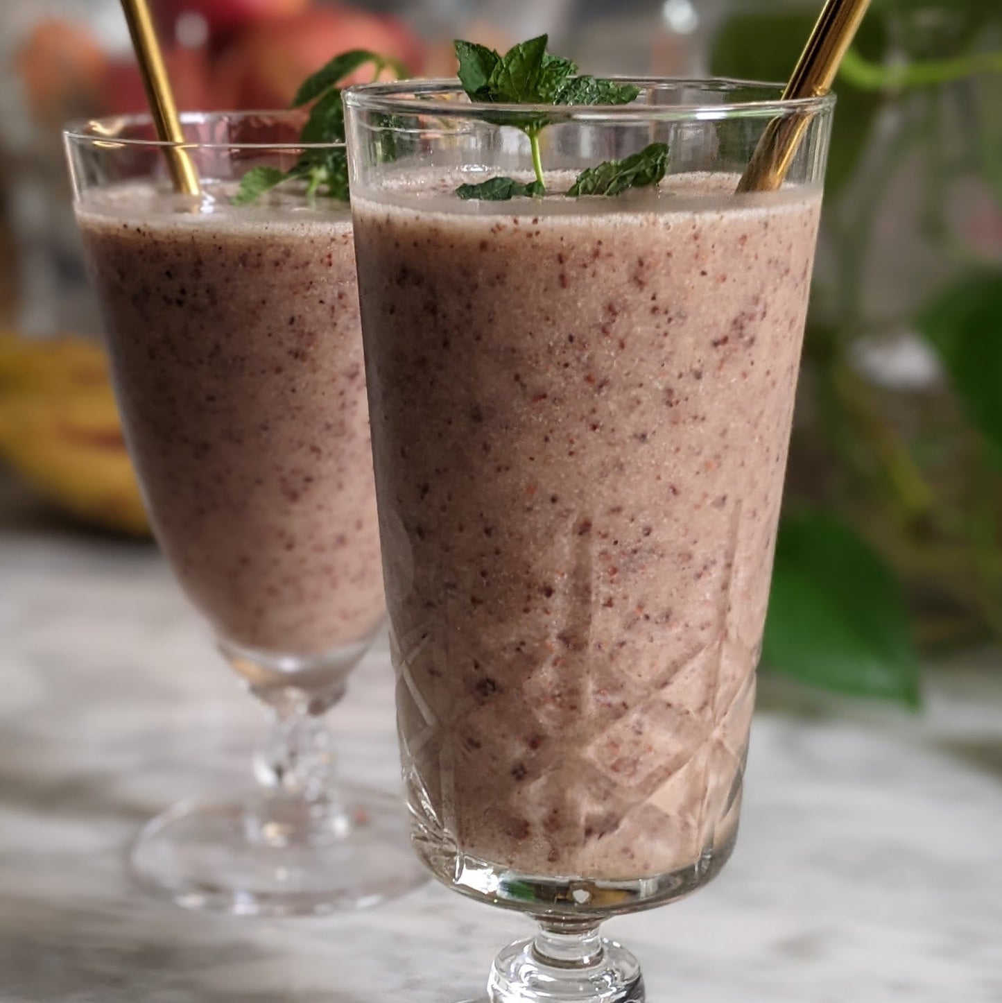 2 clear glasses on a counter filled with light, chocolate brown colored smoothies with fresh mint leaves and straws.