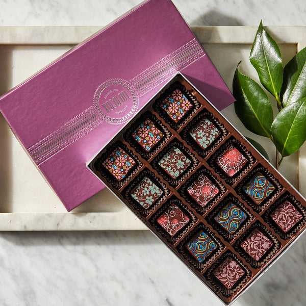 A box of various chocolate bonbons with decorative patterns on the chocolates, presented in a pink box with a leafy branch beside it.
