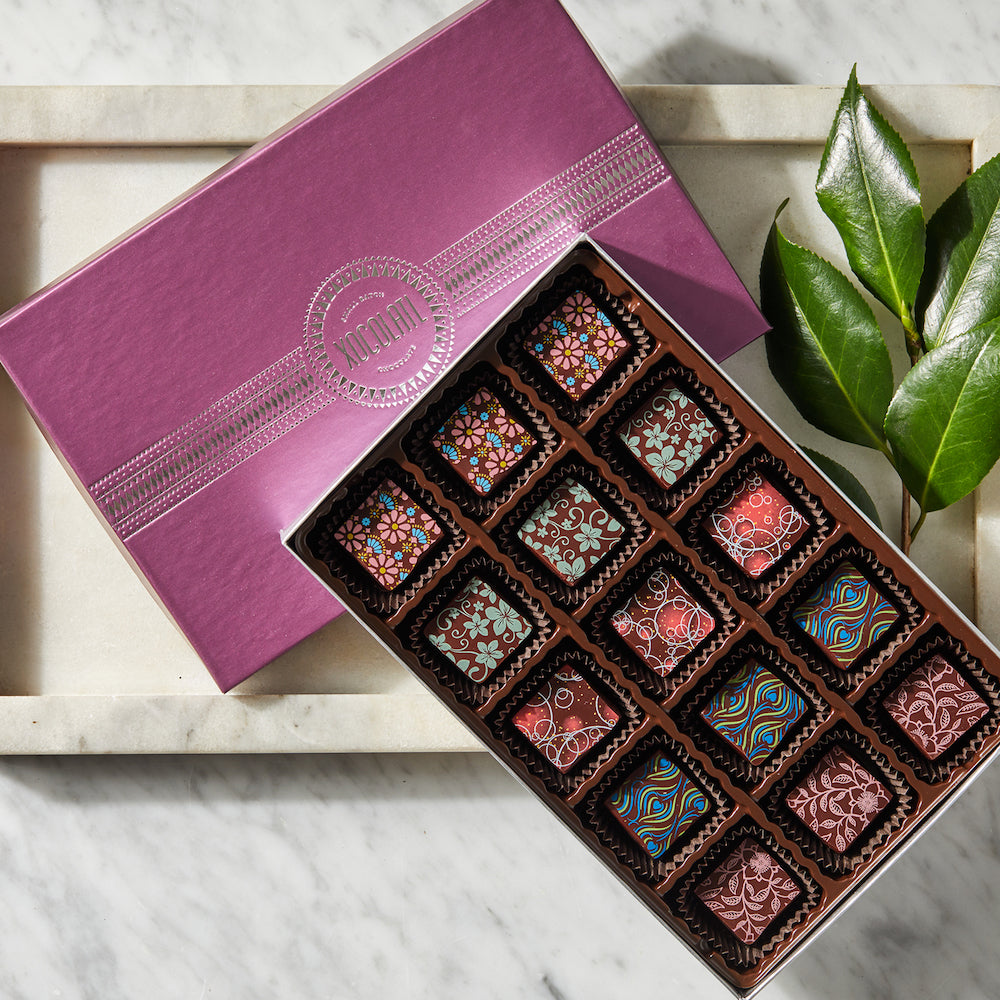 Box of chocolates with decorative patterns on a marble surface, accompanied by a purple box.