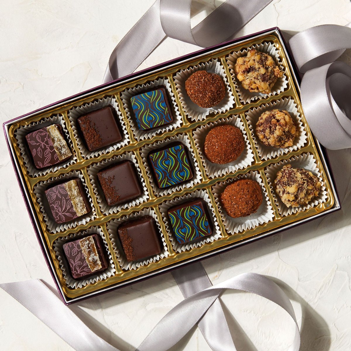 An open box of chocolate bonbons with 15  bonbons nestled inside. There are 5 flavors, each with 3 pieces. Each of the 5 flavors is indicated by a different shape & color.