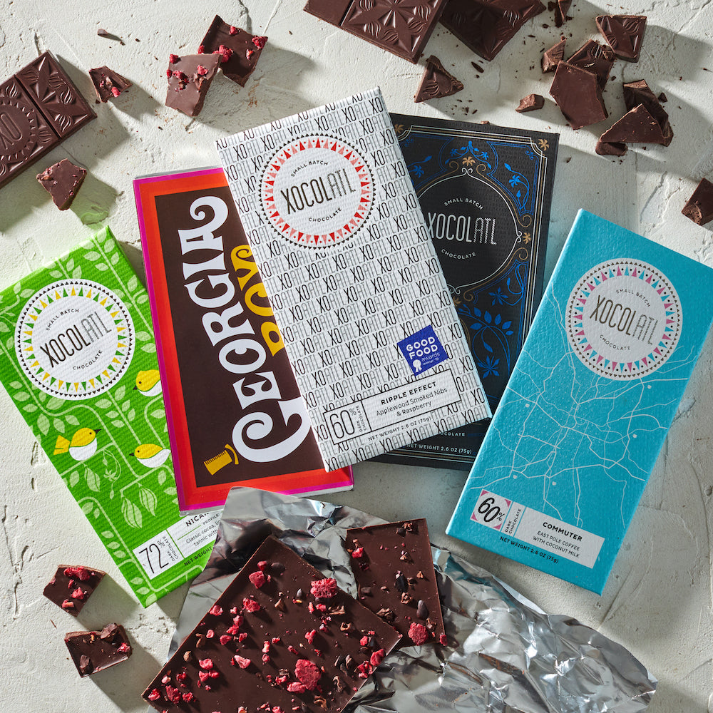 5 colorfully wrapped Xocolatl chocolate bars including flavors like "Georgia Boy" and "Commuter" along with pieces of dark chocolate fanned out on a white table.