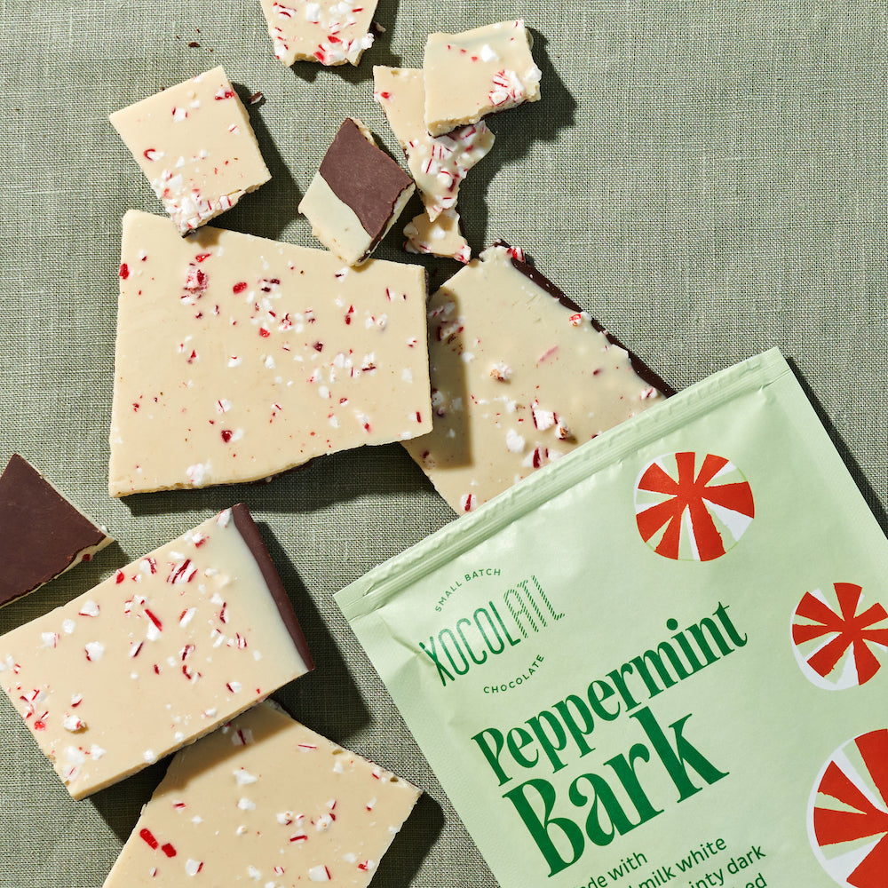 Pieces of white chocolate and dark chocolate peppermint bark topped with crushed peppermint candies spilling out of its packaging onto a green tablecloth.