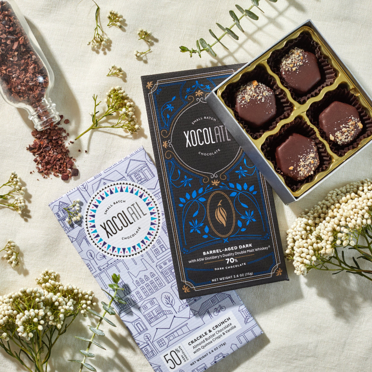 2 wrapped Xocolatl Chocolate bars in the flavors of "Crackle & Crunch" and "Barrel-Aged Dark 70%" with an open 4 piece box of decorated chocolate bonbons and a glass vial of cocoa nibs all set on a white table with small white flowers.