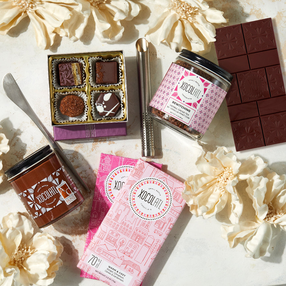 Assorted chocolate products including bars, truffles, and containers on a light surface with flowers.