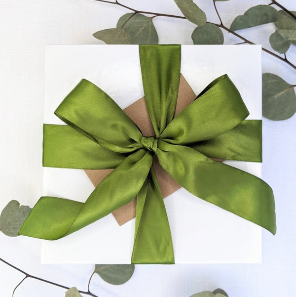 A white gift box tied with a wide, satin green bow with a note card tucked under, all on a white background with green leaves.