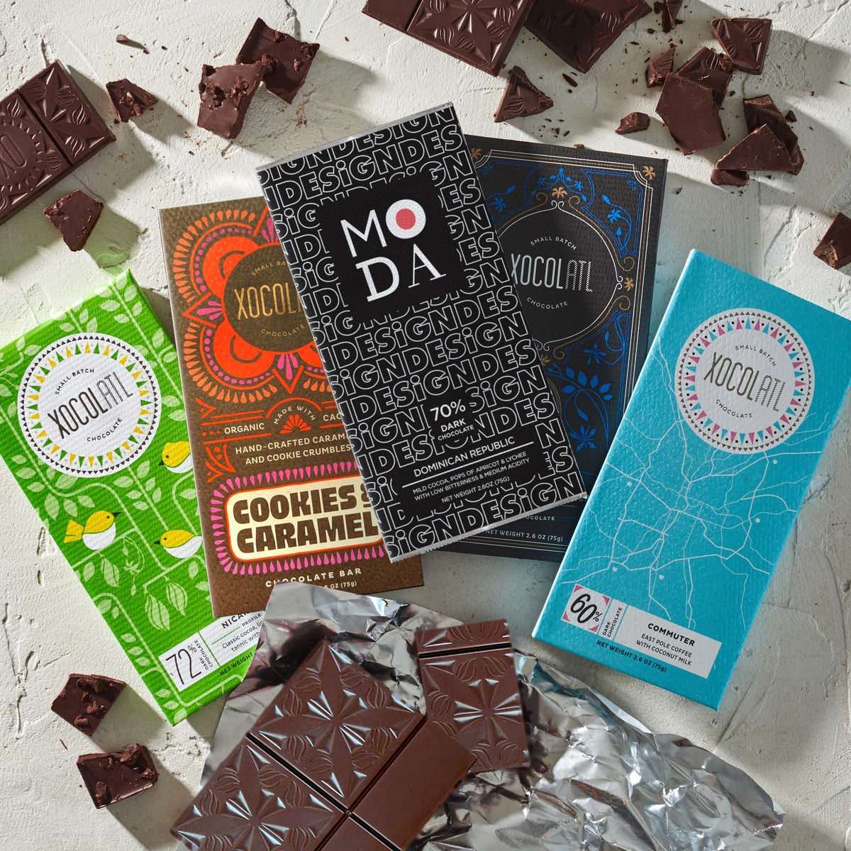 Five Xocolatl chocolate bars fanned out on a white plaster surface: Nicaragua 72% in a green bird-print wrapper, Cookies & Caramel in a brown and orange wrapper, the black-and-white MODA Dominican Republic 70%, Barrel-Aged Dark in navy blue, and Commuter in light blue, with an unwrapped bar on silver foil and broken chocolate pieces around them.