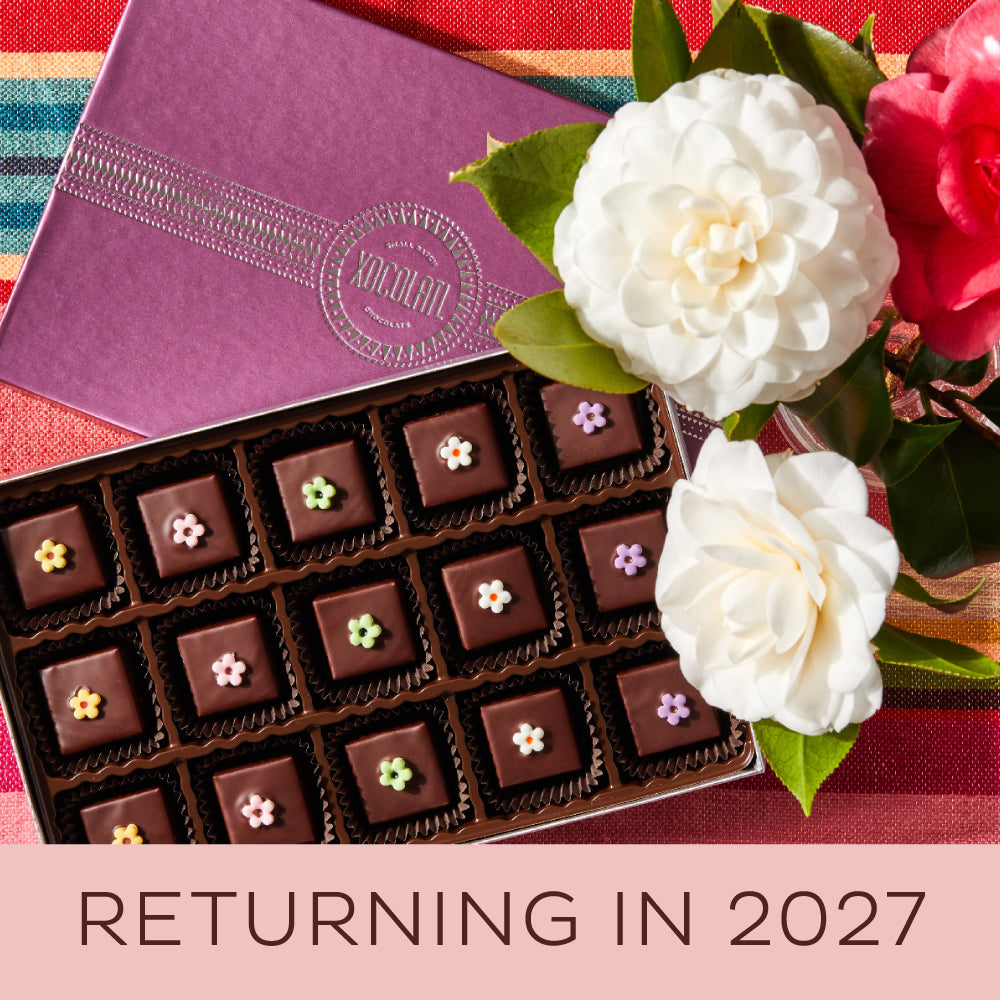 An opened box showing 15 chocolate bonbons, each topped with a colorful fondant flower, set beside white and red blooming flowers with the Xocolatl logo showing on the box top.