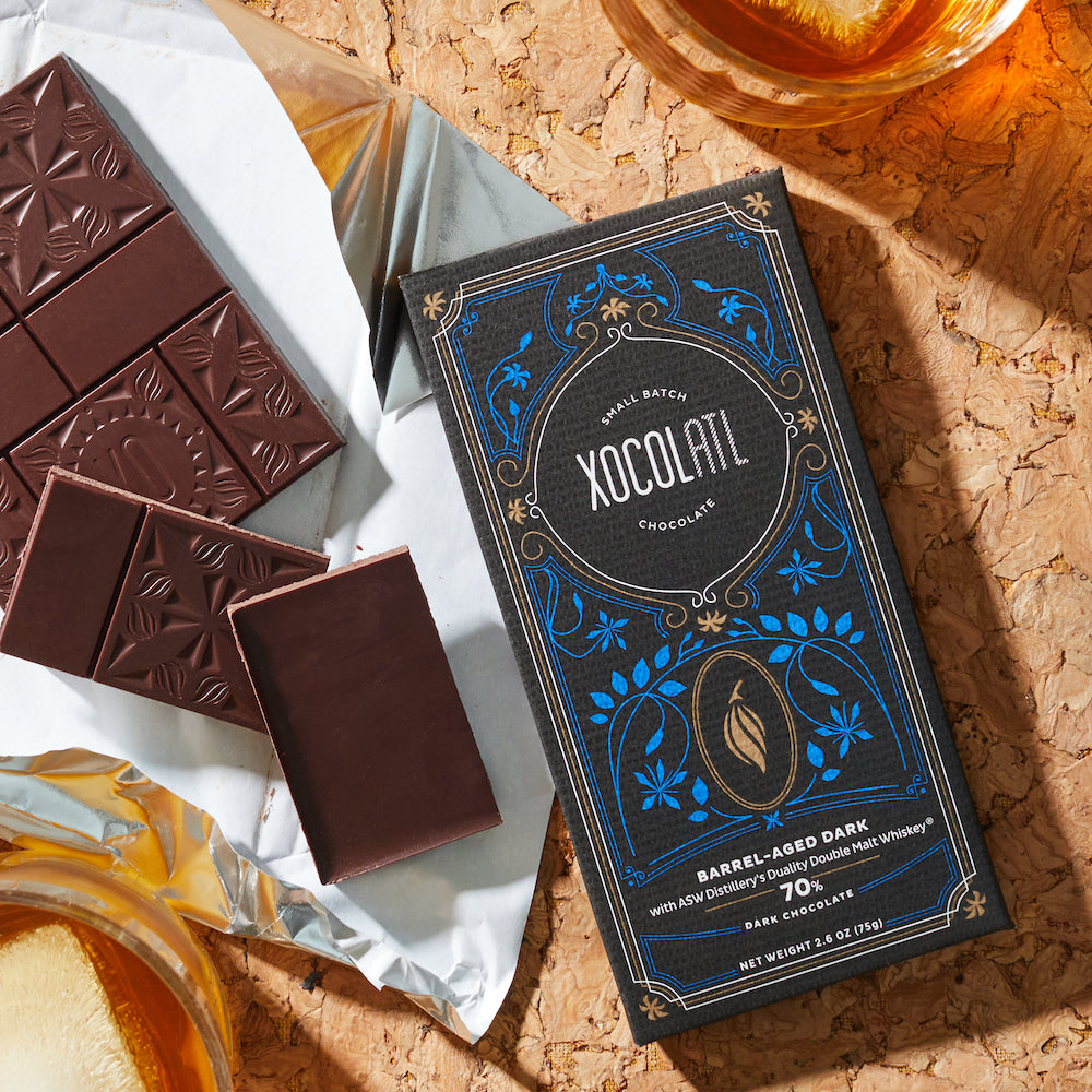 A black & blue paper wrapped Xocolatl chocolate bar of the flavor "Barrel-Aged Dark" lying beside an unwrapped bar of the same flavor, broken into pieces for imminent eating. A tumbler of whiskey is alongside.