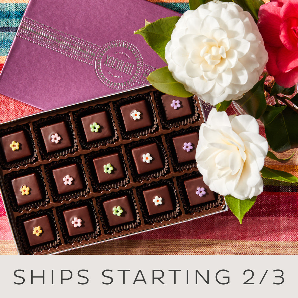 An open 15 piece box of chocolate bonbons, each topped with a colorful fondant flower and the words: Ships Starting February third.