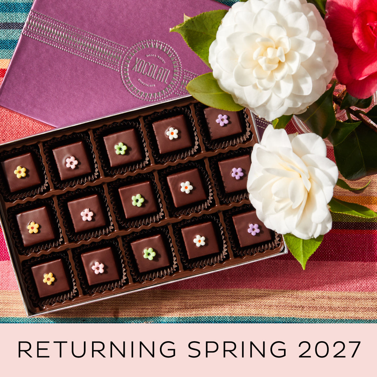 An opened box showing 15 chocolate bonbons, each topped with a colorful fondant flower, set beside white and red blooming flowers with the Xocolatl logo showing on the box top. A message at the bottom reads "Returning Spring 2027"