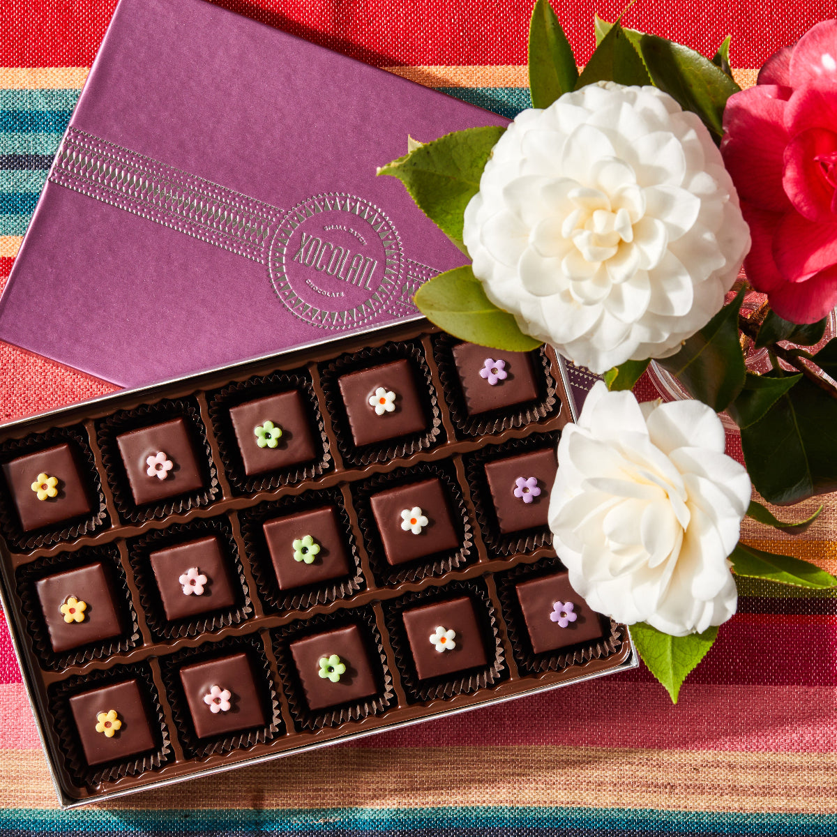 An opened box showing 15 square chocolate bonbons, each topped with a colorful fondant flower, set beside white and red blooming flowers with the Xocolatl logo showing on the box top.