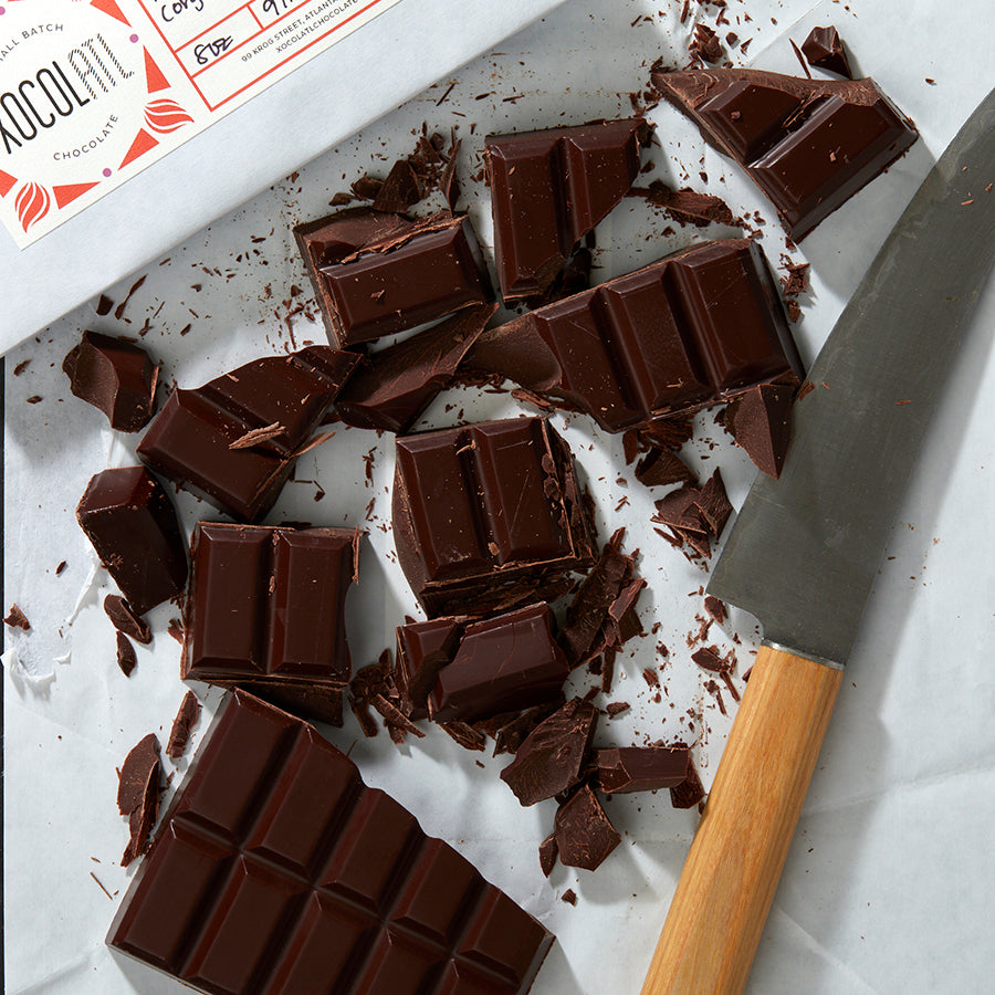 A slab of baking chocolate made up of smaller, thumb sized rectangles is roughly chopped on a white countertop surface with a chopping knife lying in the splinters & chunks of cut chocolate.