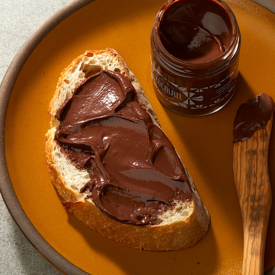 A slice of sourdough bread thickly coated with creamy chocolate hazelnut spread & ready to be eaten,  placed on a round orange plate with the jar of spread and a wooden spreading knife.