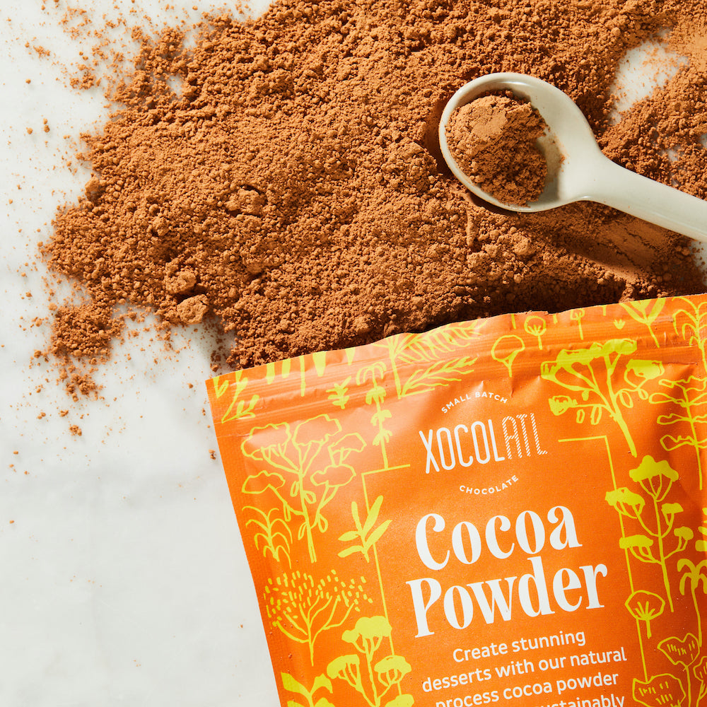 Light brown cocoa powder has spilled out of an orange and yellow pouch of Xocolatl Chocolate's Cocoa Powder, onto a white marble surface.