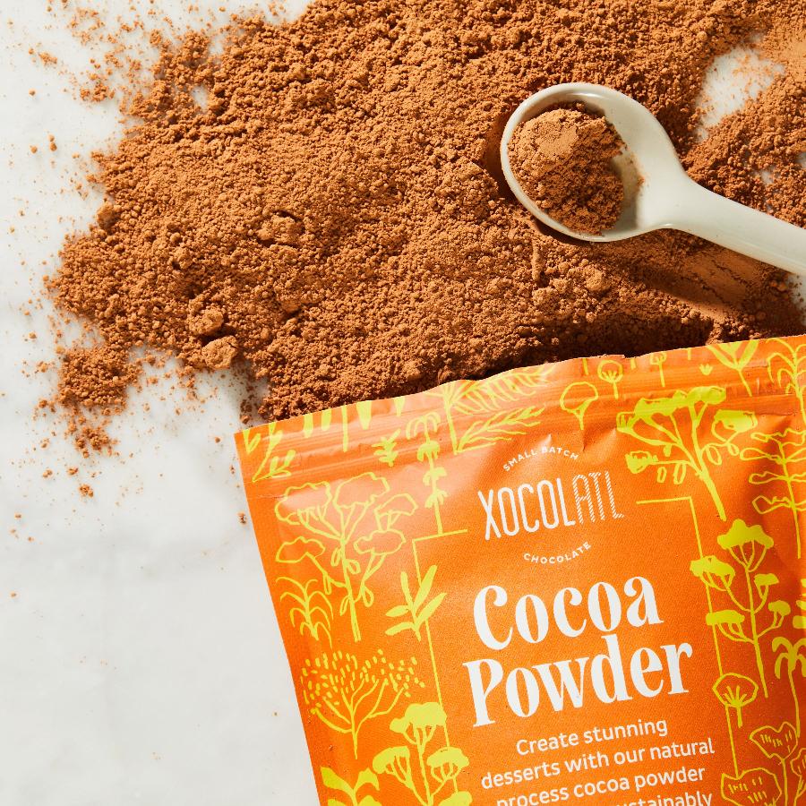 An orange, paper pouch reading, "Xocolatl Chocolate Cocoa Powder" is torn open at the top and light brown, fluffy cocoa powder has been shaken out onto a white surface with a white measuring spoon nearby.