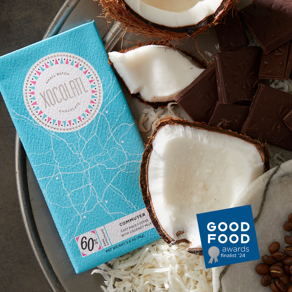 A light blue wrapped Xocolatl chocolate bar of the "Commuter" flavor, lying on a silver platter beside a fresh coconut cracked open, with a pile of chocolate squares & coffee beans. There is a blue "Good Food Awards Finalist 2024" badge on the photo.