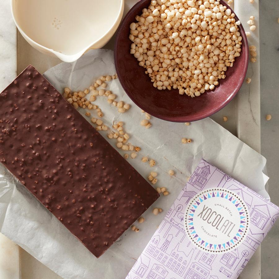 An unwrapped Xocolatl chocolate bar of the flavor "Crackle & Crunch", showing the bumpy texture of popped quinoa inside the chocolate. Also, a bowl of popped quinoa, a mug of almond milk & a lavender-wrapped Crackle & Crunch bar.