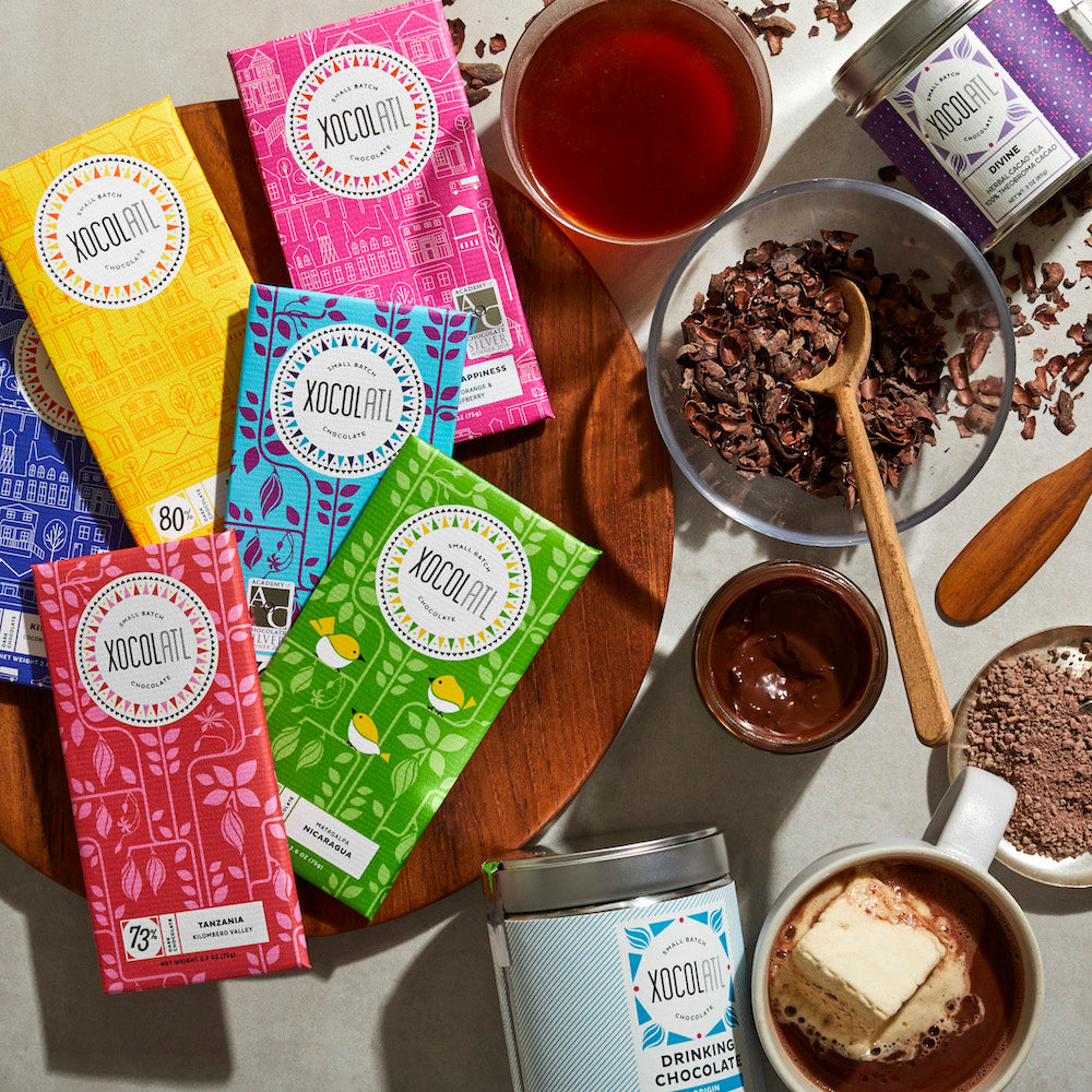 A colorful & bright display of Xocolatl chocolate bars arrayed on a wooden tray, alongside a bowl of cacao tea husks, an open jar of chocolate hazelnut spread & mugs of hot chocolate & cacao tea.