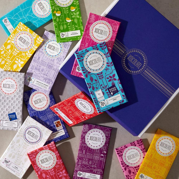 A colorful scattering of unopened Xocolatl Chocolate bars, wrapped in blues, pinks, reds, yellows, purples, whites and green. The Xocolatl bars lay beside a royal blue Xocolatl Chocolate branded gift box.