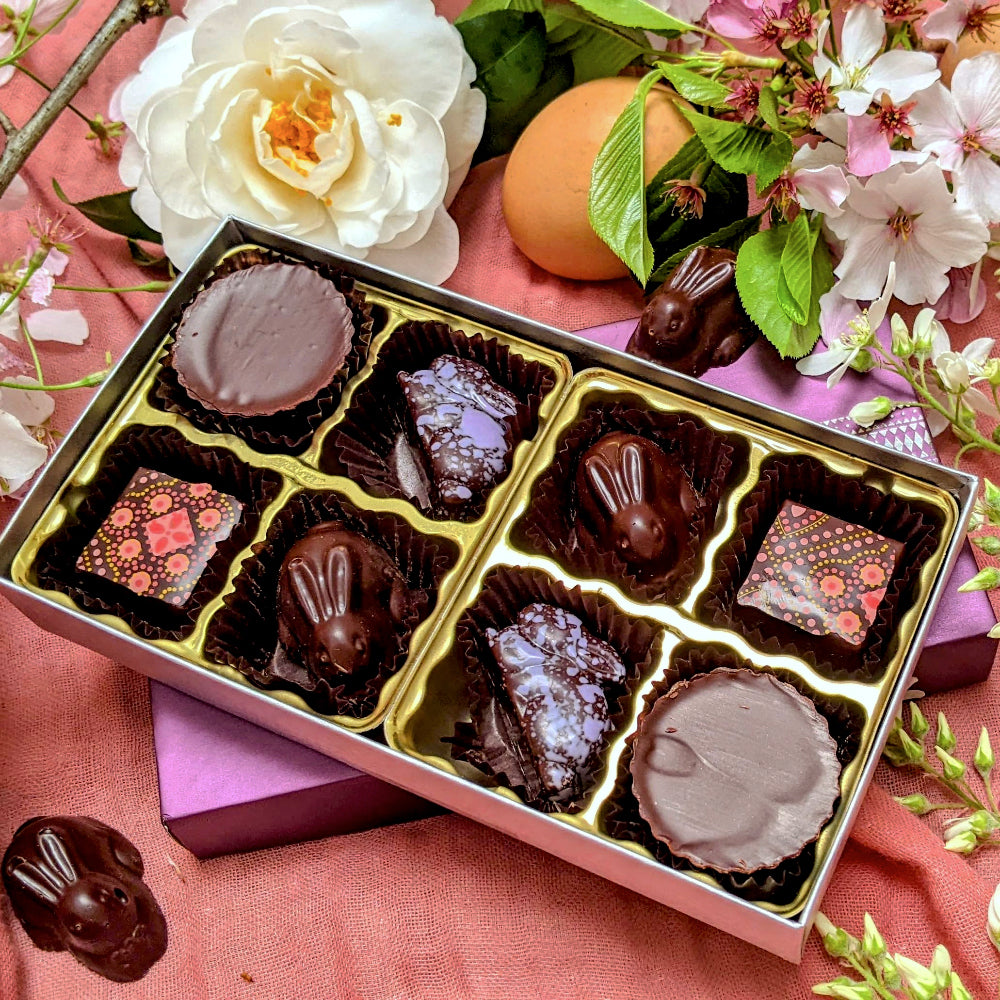An 8 piece box of chocolate bonbons opened to show a variety of bunny-shaped bonbons sitting with white and pink spring flowers.