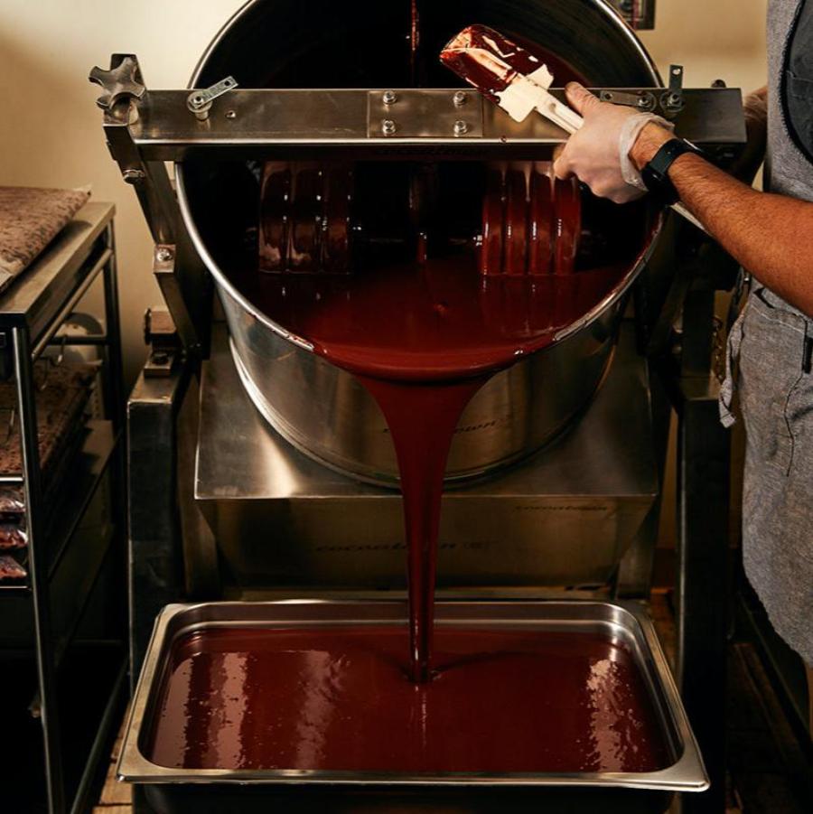 An aproned Xocolatl chocolate maker tips a stainless steel drum full of liquid chocolate down, pouring out a thick stream of chocolate into a metal pan.