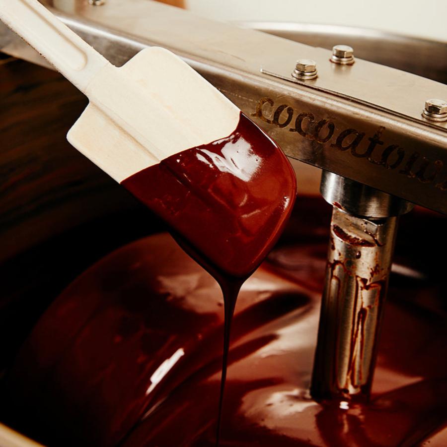 Dark brown, liquid chocolate drips off of a white rubber spatula back into a chocolate grinder full of liquid chocolate.