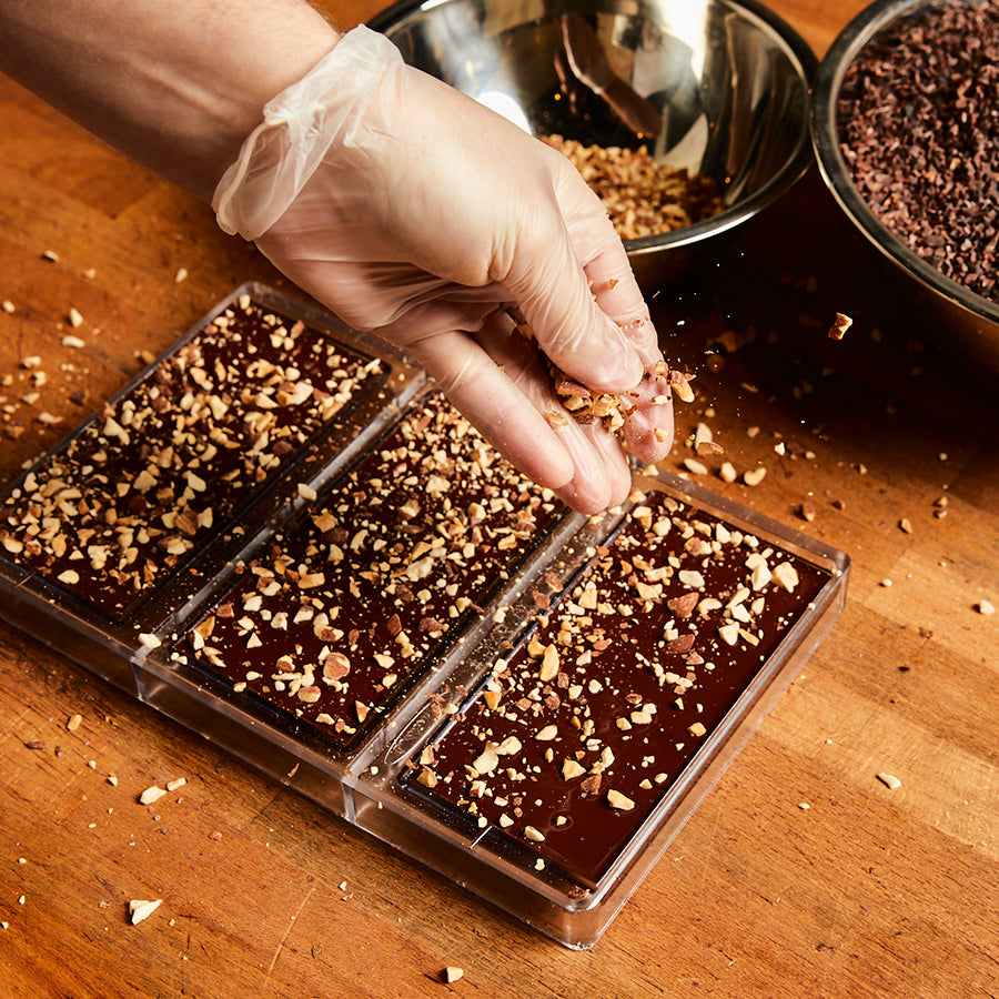 A gloved hand sprinkles roasted almond pieces onto the backs of 3 chocolate bars, sitting on a wooden chef's table.