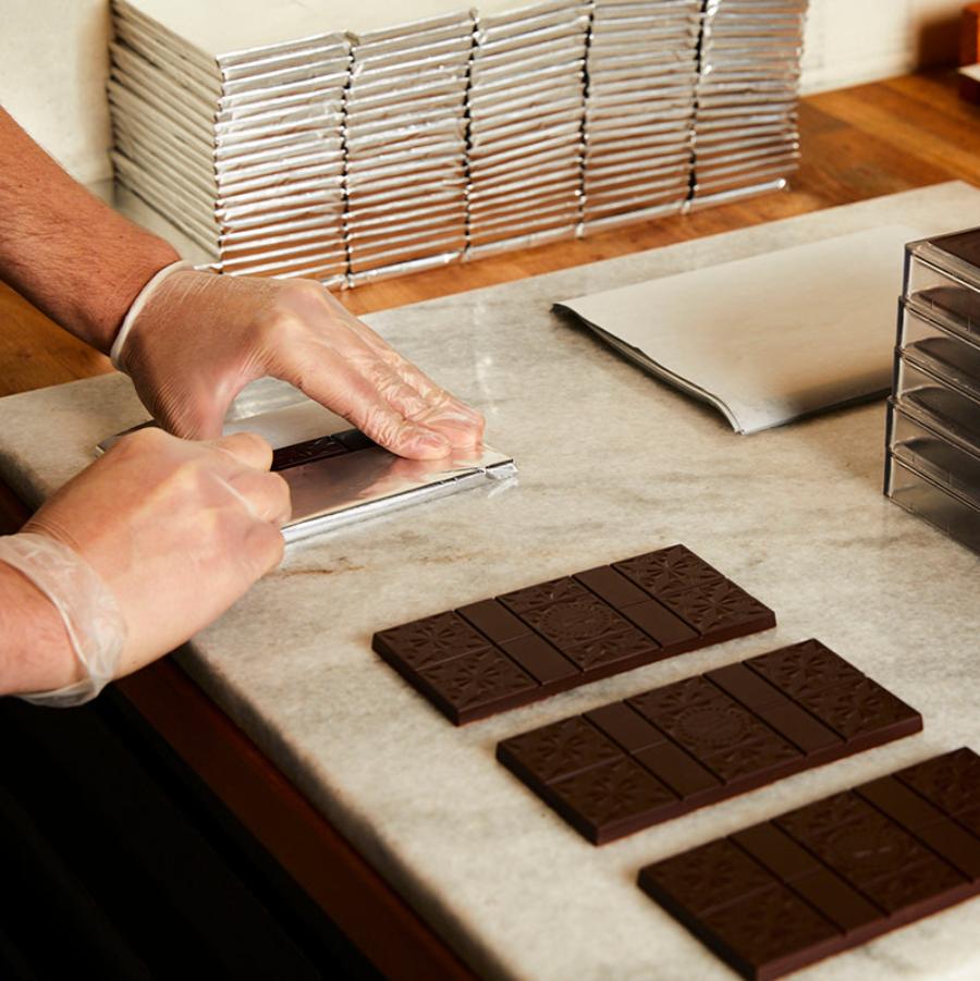 Gloved hands wrap silver foil around a Xocolatl Chocolate bar on a marble surface. In the foreground are 3 more bars to be wrapped. In the background, there are stacks of silver foil wrapped bars.