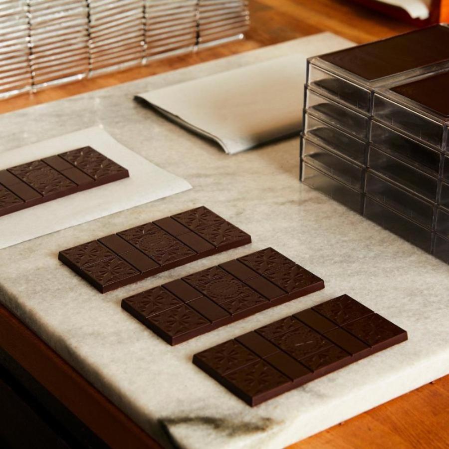 4 unwrapped Xocolatl Chocolate bars are lying on a white marble slab, waiting to be foil wrapped. A stack of silver foil bars and a stack of unwrapped bars are in the background.