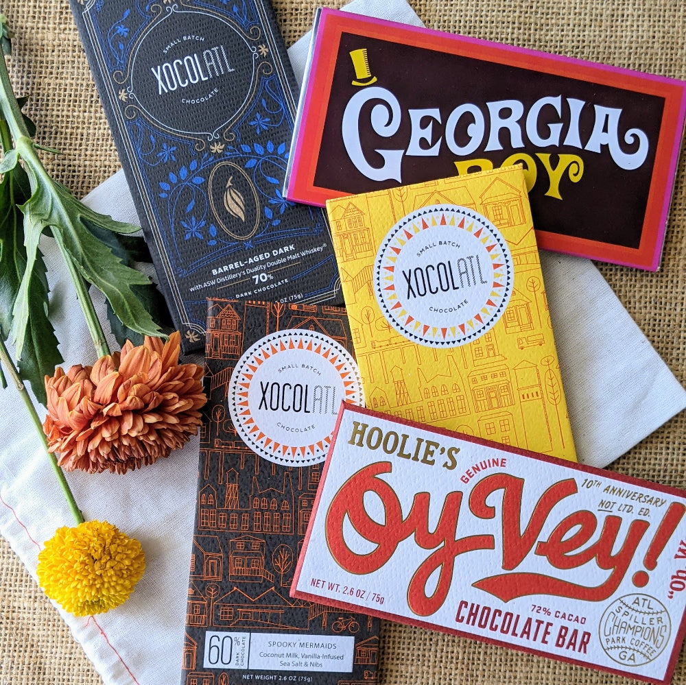 5 Xocolatl Chocolate bars on a burlap bag background with fall flowers. Flavors are Barrel-Aged Dark, Dark and Salty, Spooky Mermaids, Oy Vey and Georgia Boy.