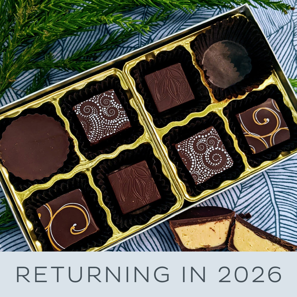 An open box showing 8 chocolate covered bonbons on a blue and white patterned picnic cloth. One bonbon is cut open showing peanut butter ganache on the inside.