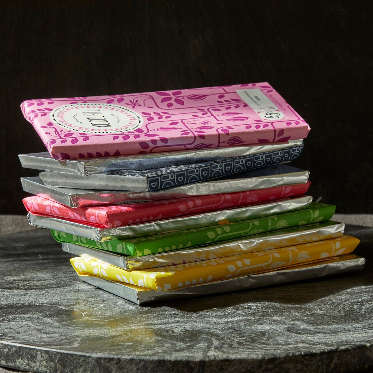5 colorfully wrapped Xocolatl chocolate bars in the colors of bright yellow, grass green, cherry red, navy blue & light pink are stacked on top of each other interspersed with bars wrapped only in silver foil.
