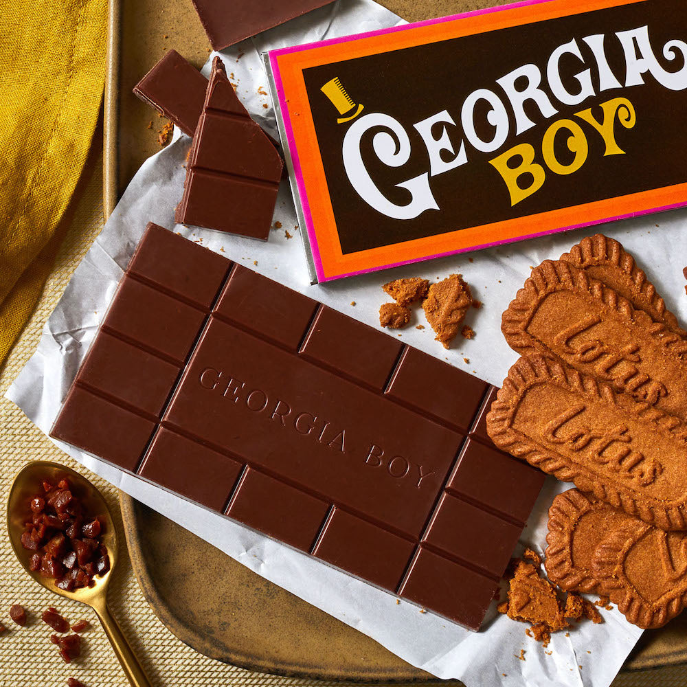 An unwrapped Xocolatl chocolate bar of the flavor "Georgia Boy", lying on a tray with opened Biscoff cookies, a spoonful of chopped caramel chunks and the bar wrapper that whimsically reads, "Georgia Boy".