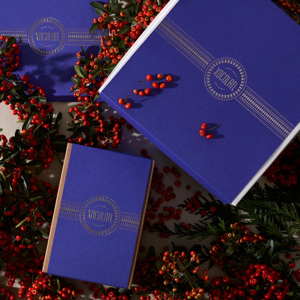 Three royal blue gift boxes (small, medium & large) with the Xocolatl logo stamped in silver foil are spread on a table covered in festive red berries & greenery.