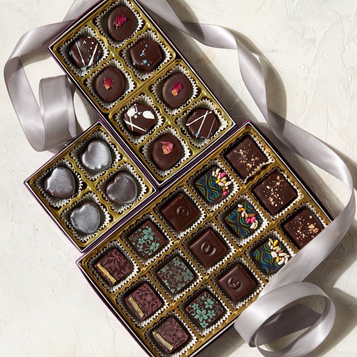 3 opened boxes full of colorfully decorated chocolate bonbons are displayed on a white background with a silver satin ribbon twining around the boxes. The large box has 15 chocolates, the medium box has 8 chocolates & the small box has 4 chocolates, all variously decorated.