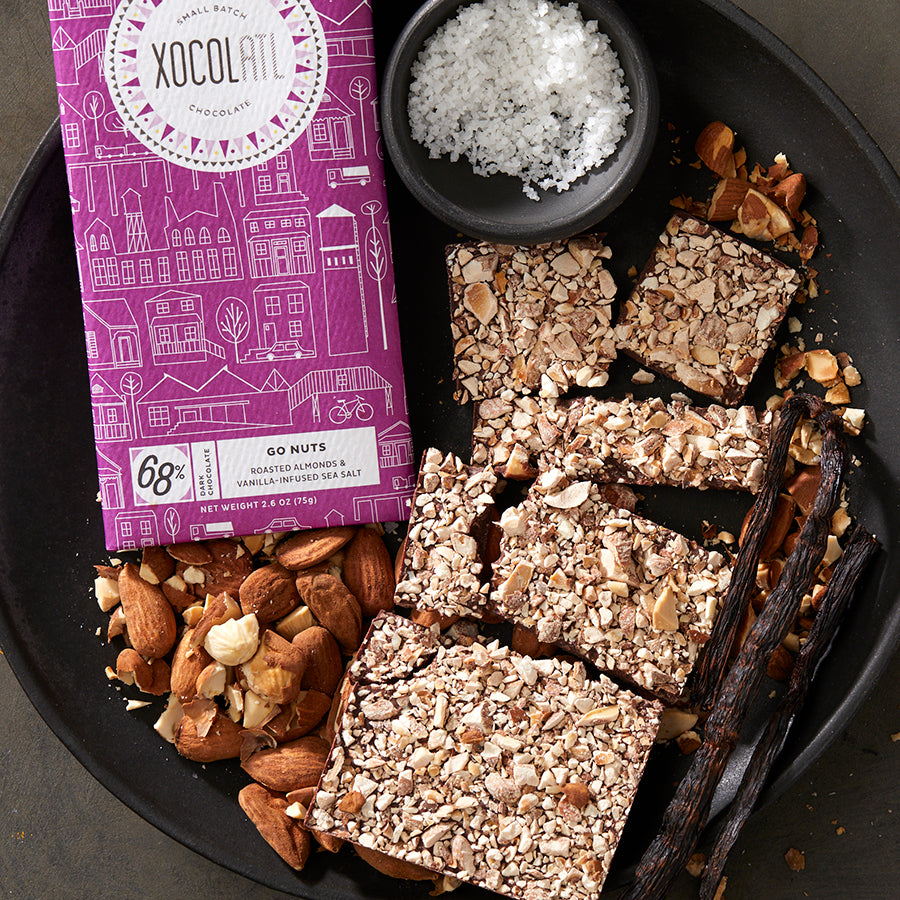 A purple wrapped Xocolatl chocolate bar of the flavor "Go Nuts" and an unwrapped chocolate bar showing a covering of crushed almonds, on a black plate with sea salt, almonds and vanilla pods.