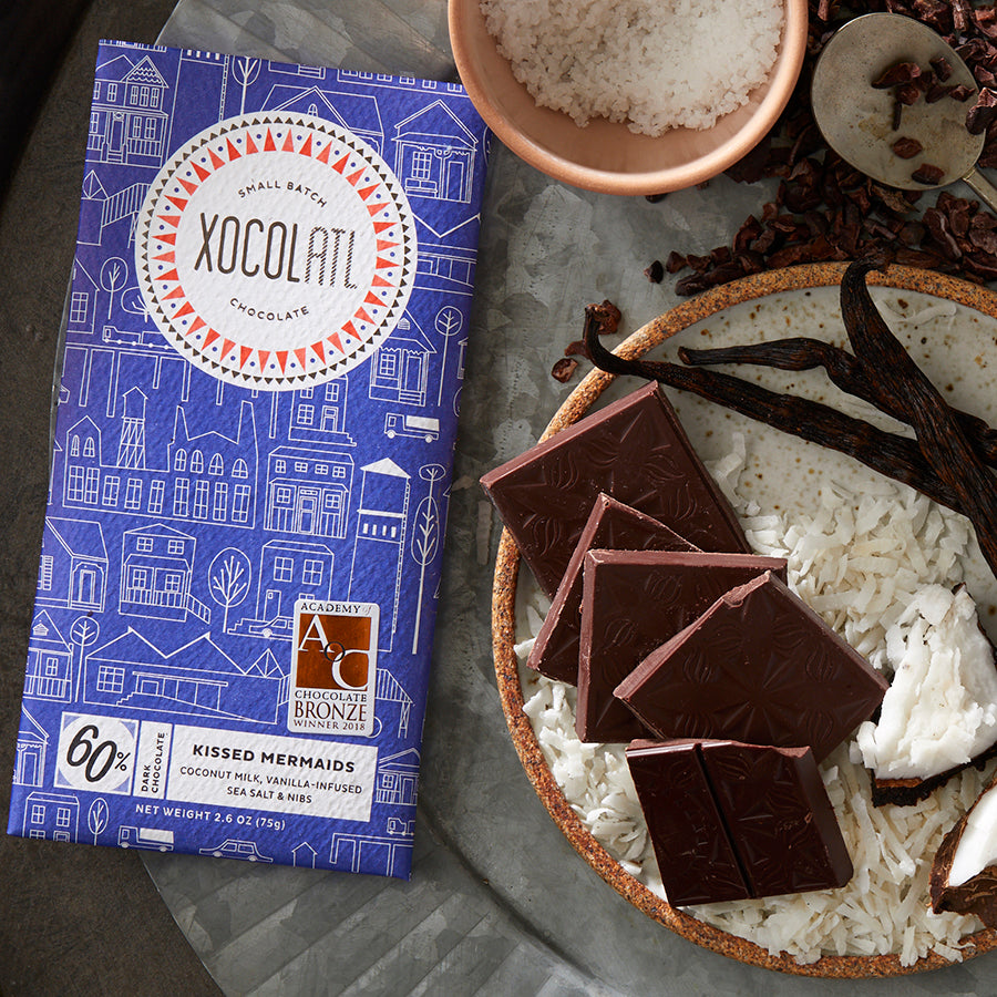 A blue-wrapped "Kissed Mermaids" flavored Xocolatl chocolate bar beside squares of this chocolate bar & bowls filled with coconut shreds, sea salt, vanilla beans & cacao nibs.