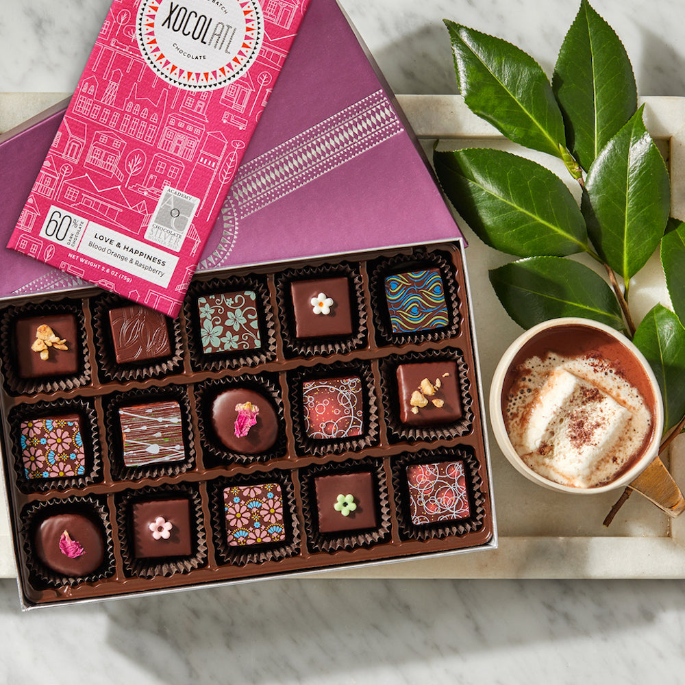 An open 15 piece box of chocolate bonbons, variously and colorfully decorated, with a pink wrapped Xocolatl Chocolate bar and a cup of hot chocolate with a melting marshmallow, all on a white marble tray.