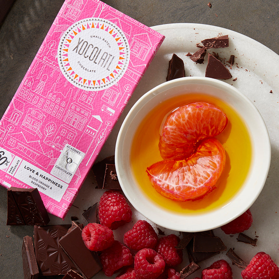 A hot pink wrapped Xocolatl chocolate bar of the flavor "Love & Happiness" beside bowls filled with chocolate squares, fresh raspberries & blood orange segments in olive oil.