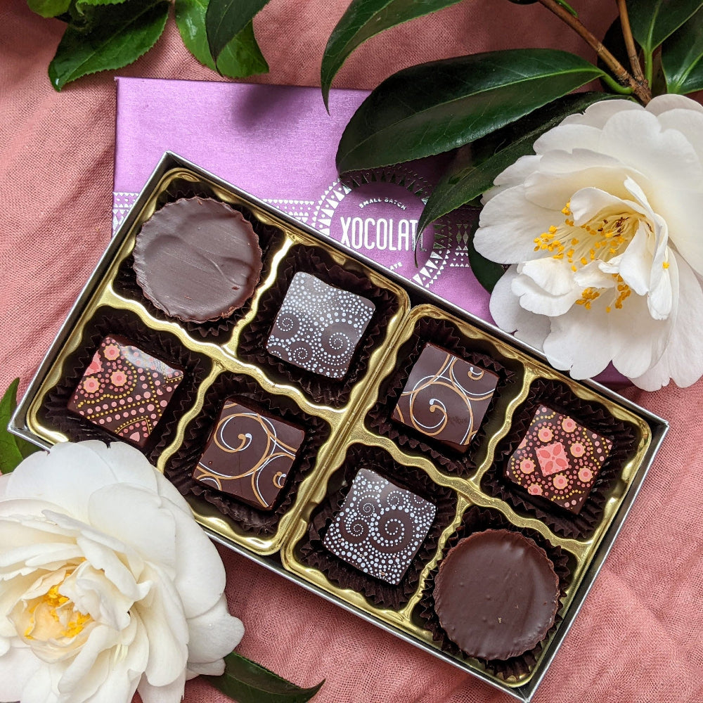 An open 8 piece box of chocolate bonbons in a variety of colors and designs, along with 2 white flowers on a pink background. 