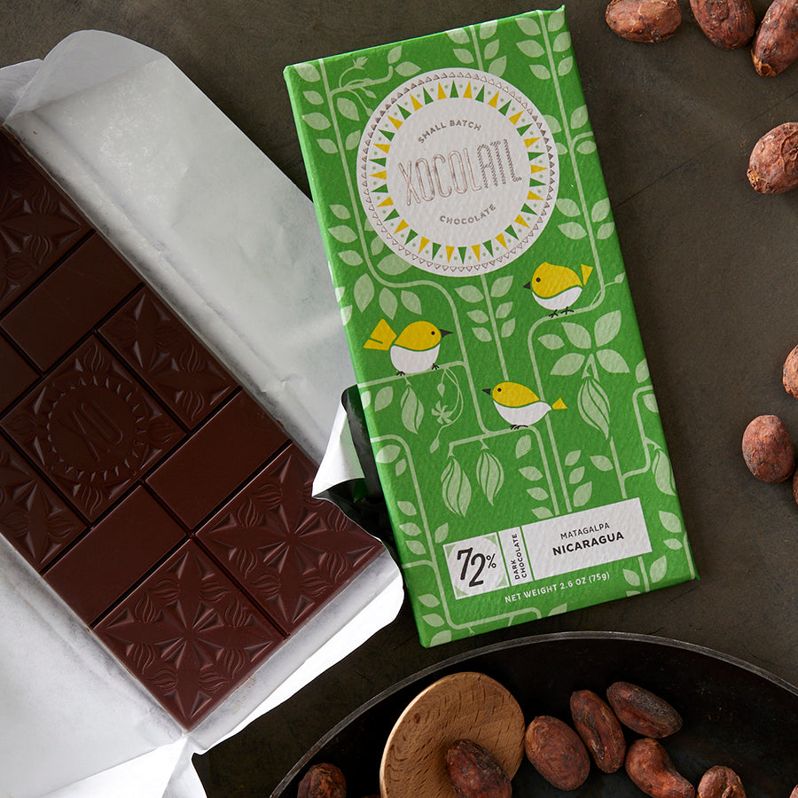 An unwrapped Xocolatl chocolate bar with   a floral design beside a wrapped "72% Single Origin Nicaragua" bar in a bright green wrapper with a pattern of yellow birds.