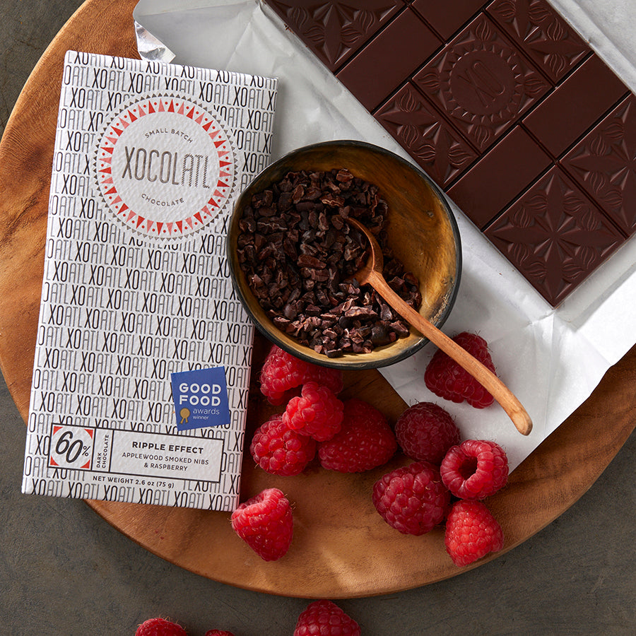 A black and white Xocolatl chocolate bar wrapper of the flavor "Ripple Effect" lying on a wooden plate with a bowl of cacao nibs, a pile of fresh raspberries & an opened chocolate bar.